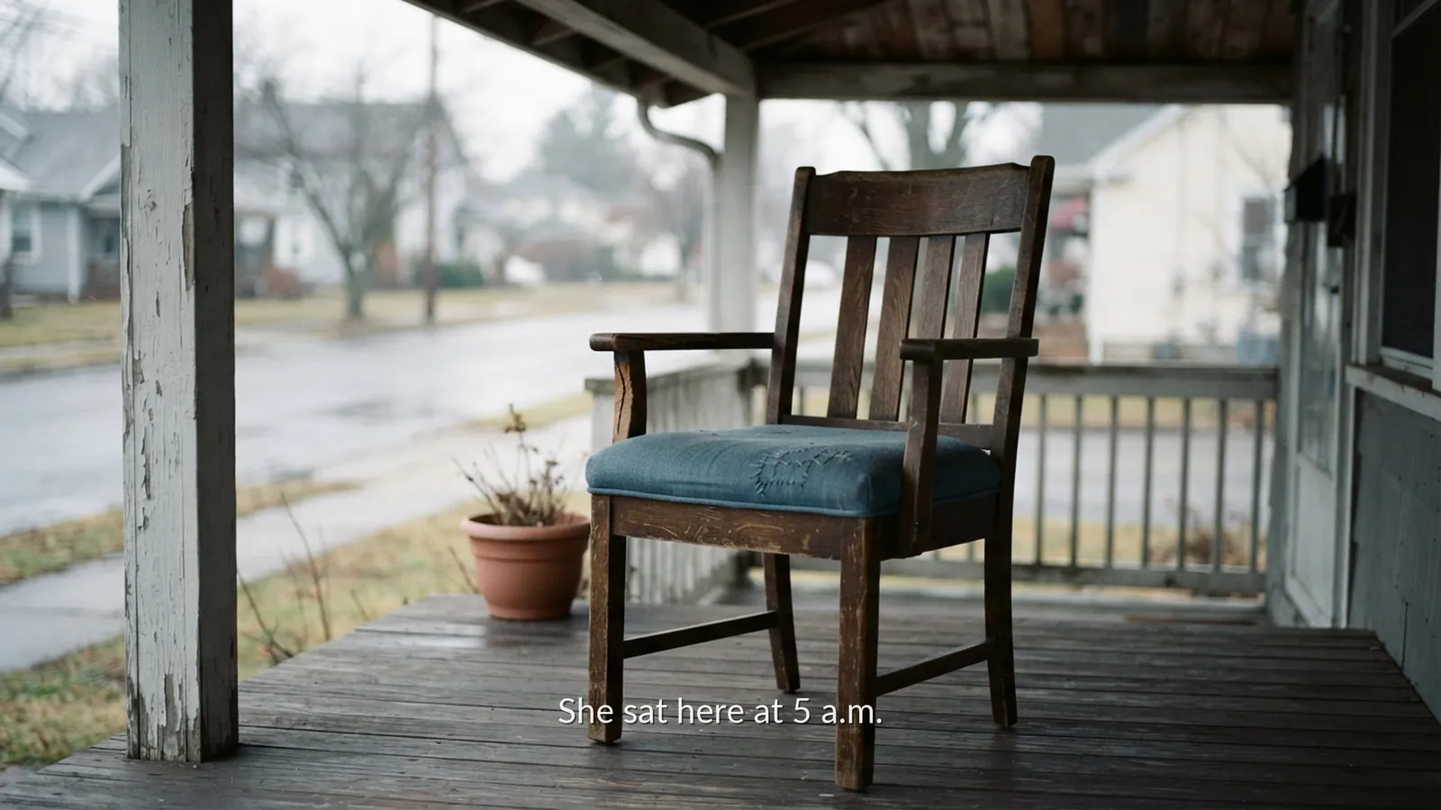 An empty wooden chair with a mended blue cushion on a porch in the rain